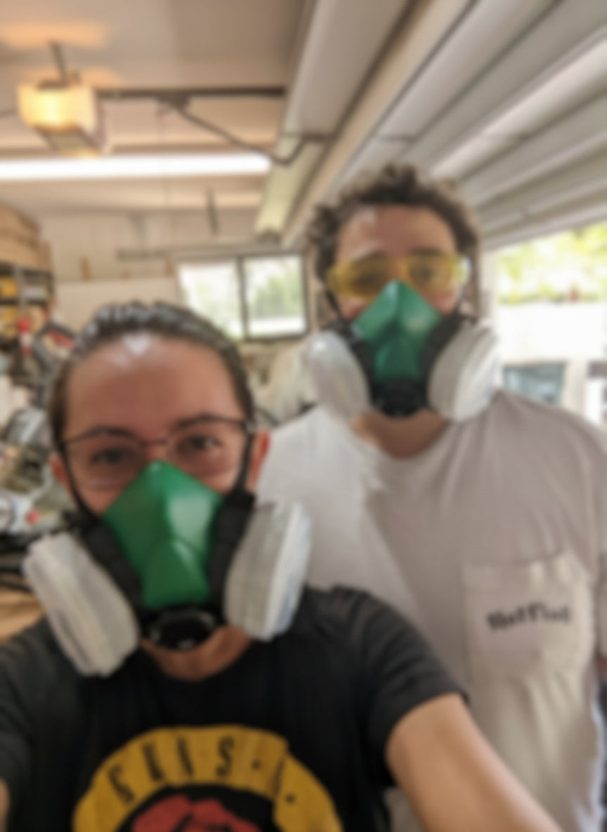 Two people wearing protective gear, filtered masks and safety goggles, in their garage woodshop